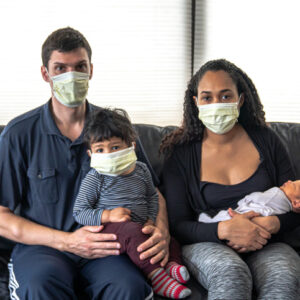 A mixed family sits on a black leather sofa with father and mother holding their infant son and daughter all wearing medical face masks in hopes of preventing getting sick from coronavirus