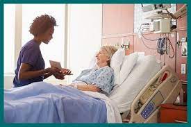 a nurse in royal blue scrubs sitting on the bed of an elderly female patient for excellent care